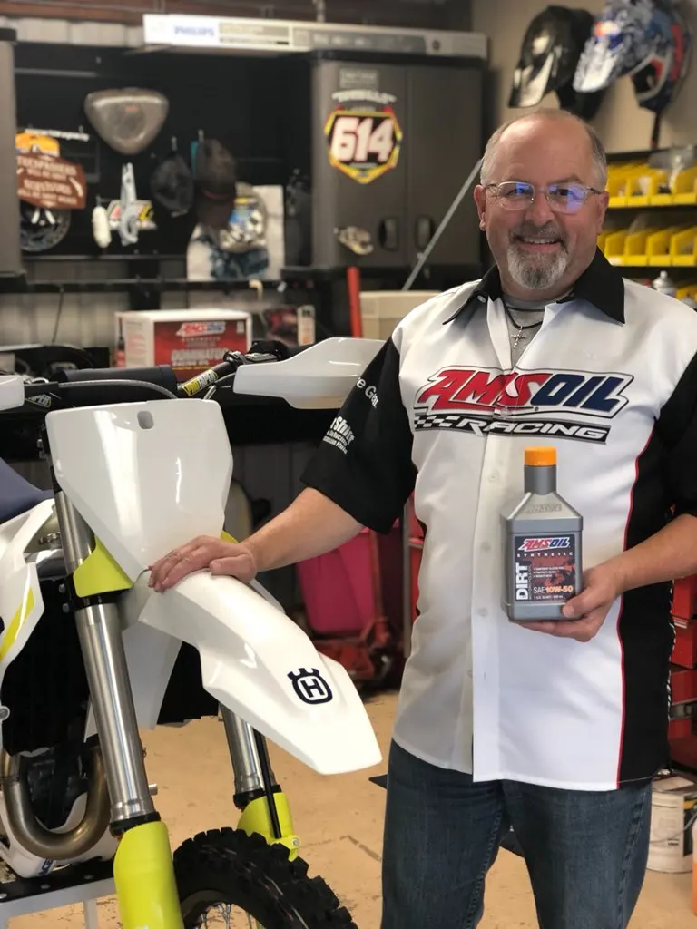Mark Ossinger in an AMSOIL Racing shirt holding AMSOIL Dirt Bike oil next to a race bike